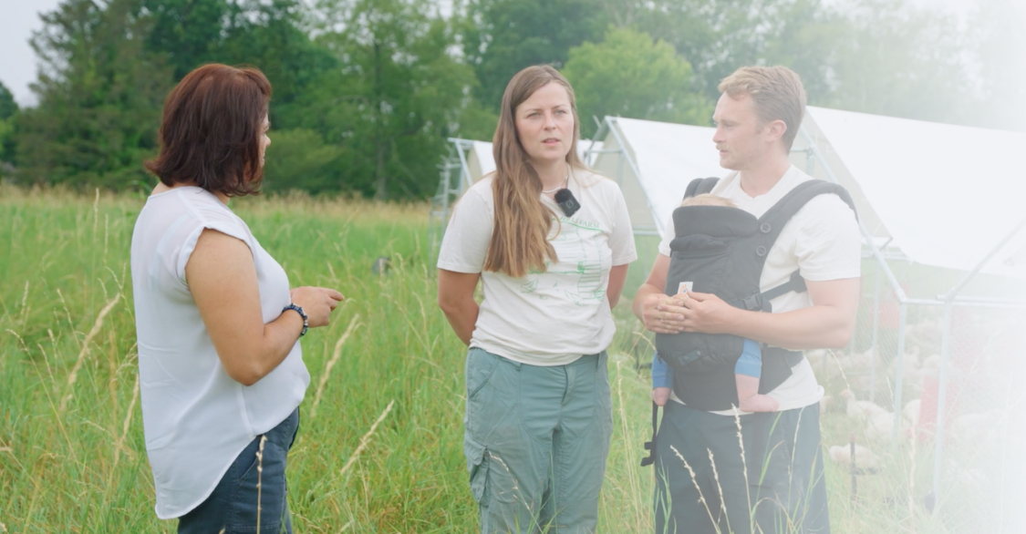 3 people talking in a field of tall grass with chicken tractors in the background